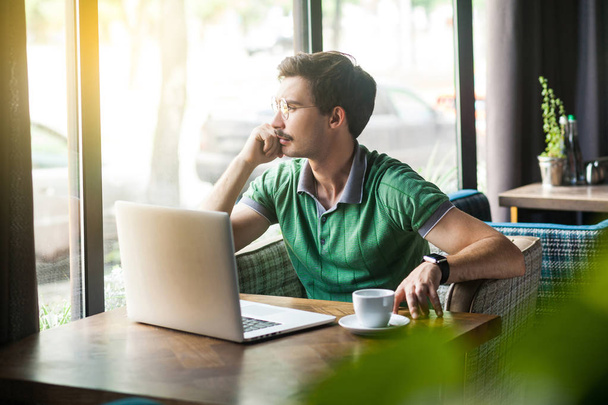 Young thoughtful businessman in green t-shirt working on laptop and looking to outside while sitting in cafe, freelancing concept - Foto, afbeelding