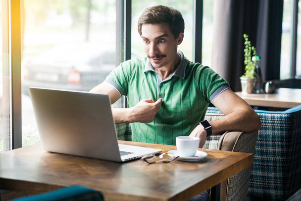 Young amazed businessman in green t-shirt pointing and looking at laptop display with surprised face while sitting in cafe, freelancing concept - Foto, Imagem