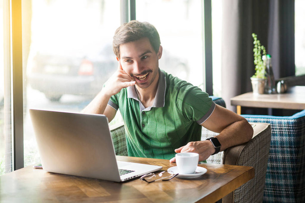 Young funny businessman in green t-shirt looking at laptop screen on video call and showing call gesture while sitting in cafe, freelancing concept - Foto, immagini