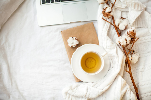Uma caneca branca com chá, algodão, um laptop, uma xadrez e um suporte de livro na cama. Pequeno-almoço na cama. Aconchegante. Outono. Inverno
. - Foto, Imagem
