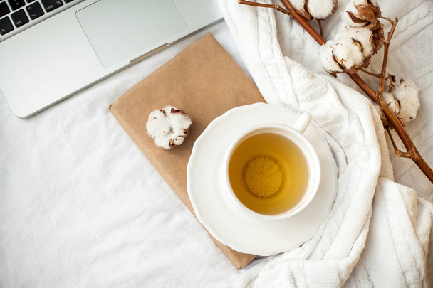 Μια άσπρη κούπα με τσάι, βαμβάκι, ένα φορητό υπολογιστή, ένα καρό και ένα βιβλίο στέκεται στο κρεβάτι. Πρωινό στο κρεβάτι. Άνετο. Φθινόπωρο. Χειμώνα. - Φωτογραφία, εικόνα