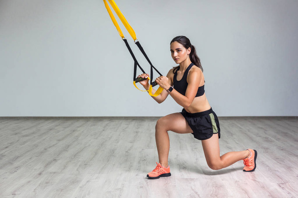  young beautiful fit woman in sportswear doing squat exercise on one knee with fitness straps in gym. Functional training concept     - Foto, immagini