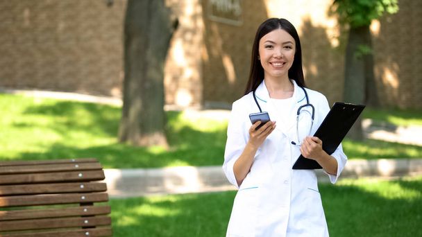 Médico sonriente sosteniendo teléfono inteligente y tableta en consulta clínica médica parque
 - Foto, Imagen