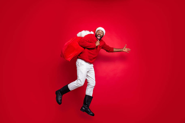 Full body profile side photo of cheerful guy in xmas santa claus hat holding carrying bag in christmas night wearing pullover pants trousers boot isolated over red background - Foto, imagen