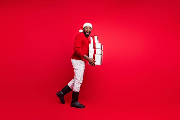 Full length photo of cheerful promoter holding packages with ribbon for christmas night wearing santa claus hat cap pants jumper isolated over red background - Photo, Image