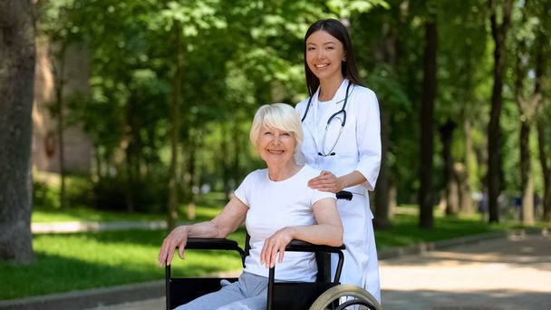 Enfermeira feminina e mulher idosa deficiente sorrindo para a câmera, relaxar no parque hospitalar
 - Foto, Imagem