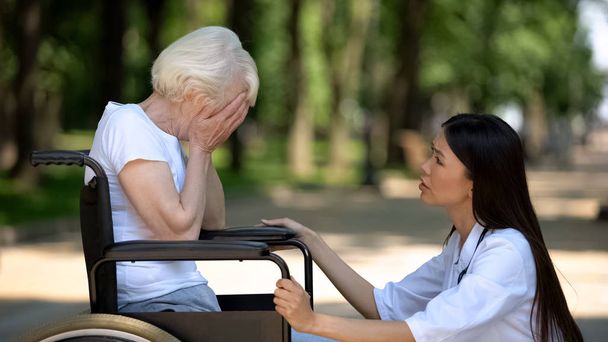 fürsorgliche Krankenschwester beruhigt weinende behinderte Seniorin im Rollstuhl, langsame Genesung - Foto, Bild