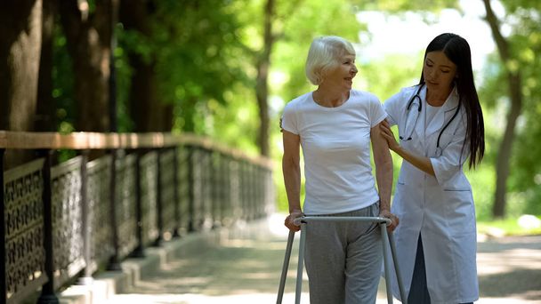 Caring nurse helping senior disabled woman with frame walk in park, rehab - Fotoğraf, Görsel
