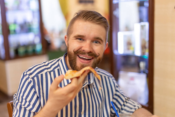 Portrait of a happy handsome young guy with a mustache and a beard eating a piece of pizza in a cafe. The concept of unhealthy fastfood. - Photo, Image