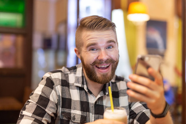 Stylish young man with a mustache and beard is watching a social network using a smartphone while sitting in a cafe on a weekend. The concept of dependence on social networks. - 写真・画像