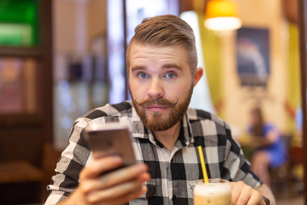 Retrato de un joven apuesto y reflexivo con bigote y barba mirando a la cámara en un café sobre un fondo borroso. El concepto de estudiante o los hombres con estilo
. - Foto, Imagen
