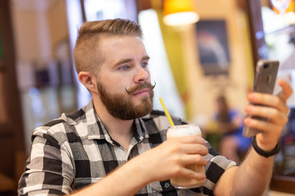 Стильний молодий чоловік з вусами і бородою дивиться соціальну мережу, використовуючи смартфон, сидячи в кафе на вихідних. Концепція залежності від соціальних мереж
. - Фото, зображення