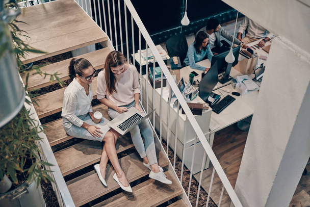 Top view of two young modern women in smart casual wear communicating and using laptop while sitting on the stairs     - Foto, Imagen