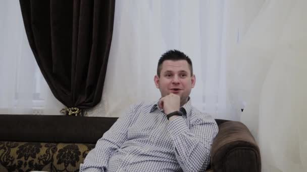 ein emotionaler Mann sitzt auf einem Sofa in einem Hochzeitssalon und reagiert auf Kleider, die seine Frau zeigt. - Filmmaterial, Video