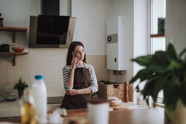 Serene lady is speaking on phone in kitchen - Фото, изображение
