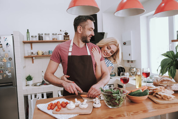 Hurmaava blondi nainen halaa miehensä takaapäin, kun hän kokkaa illallista
 - Valokuva, kuva
