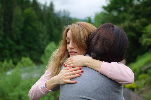 Young blonde woman is hugging her senior mother enjoying being together in the mountains during vacation. Concept of tenderness, care, family love, relationships. - Photo, Image