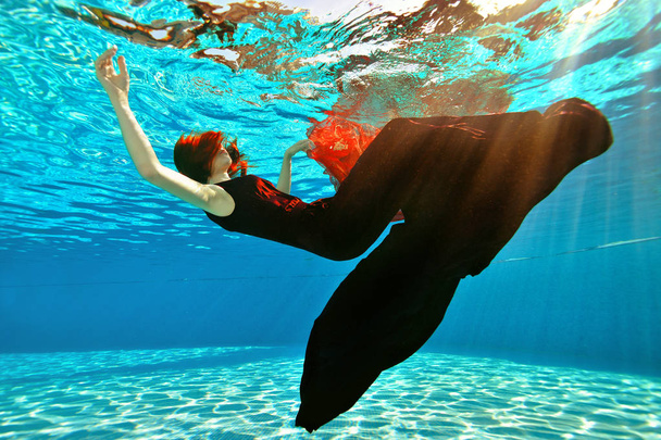 Unusual girl drowns in the pool. A girl with red hair and a dress falls to the bottom of the pool on the background of sunlight. Surrealism. Concept. - 写真・画像
