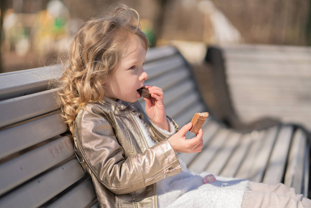 emotional portrait of a little girl with sweets, sitting on the bench in the city park outdoor alone. pretty child have a bad tasty snack. - Fotoğraf, Görsel