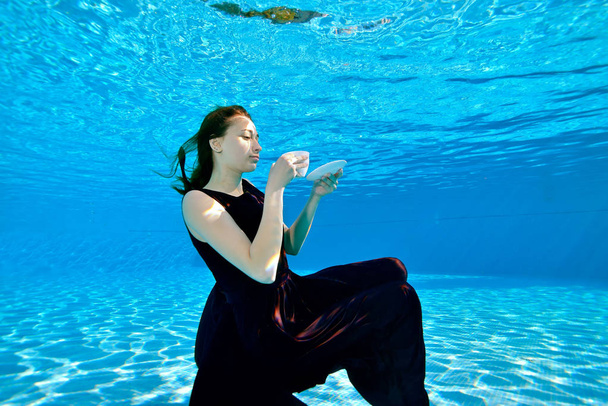 Cute young girl posing underwater at the bottom of the pool with a white Cup in her hand, in a Burgundy dress on a blue background. Tea party underwater. Surrealism. Concept. Portrait - Photo, Image