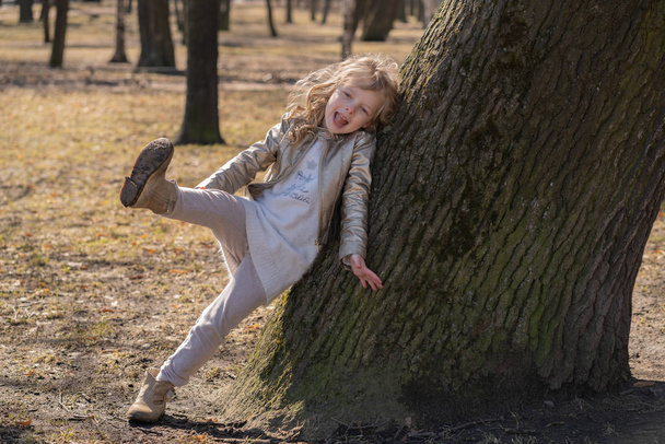 pretty curly kid girl in leather jacket posing near big tree in the city park alone - Photo, Image