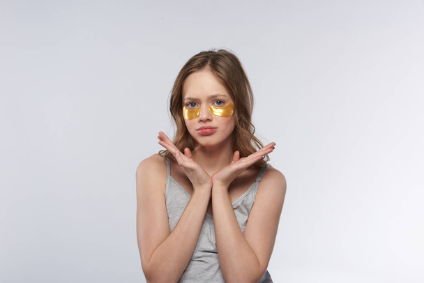 Upset teen girl in tank top using gold under-eye patches - Zdjęcie, obraz
