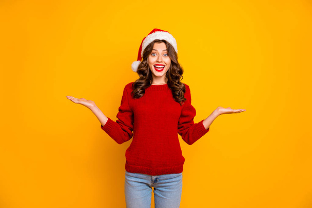 Beautiful lady proposing novelty on open arms wear red knitted sweater and santa hat isolated yellow background - Foto, Bild