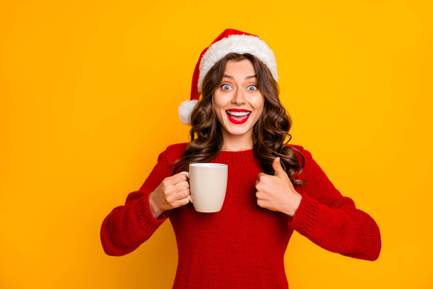 Pretty lady holding hot beverage in hands advising cool drink wear knitted jumper and santa hat isolated yellow background - Photo, image