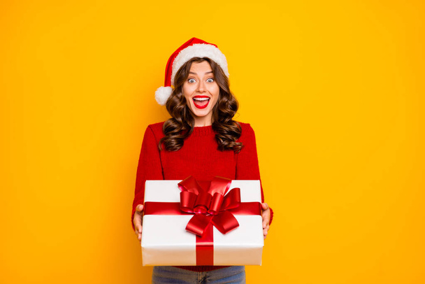 Beautiful lady holding large giftbox in hands wear knitted sweater and santa hat isolated yellow background - Zdjęcie, obraz