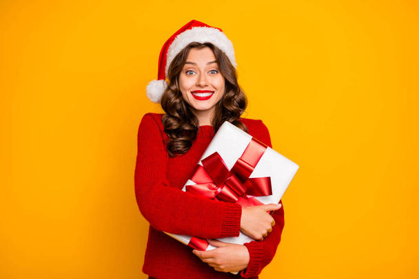 Beautiful lady holding large giftbox close to chest wear knitted jumper and santa hat isolated yellow background - Zdjęcie, obraz