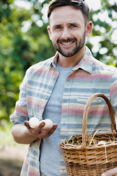 Portrait of smiling farmer holding eggs in basket at farm - Фото, изображение