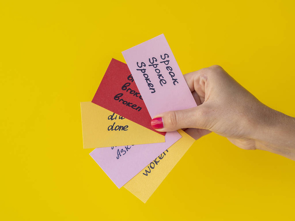 Female hand holds flashcards with handwritten irregular English verbs isolated on yellow. Language learning concept - 写真・画像