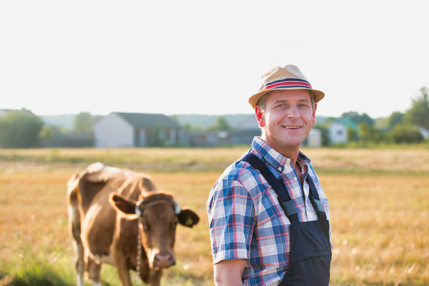 Portrait of mature farmer standing against cow on field at farm - Photo, Image