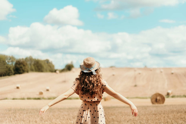 Girl with curly hair, straw hat, polka dot dress lifting her hands up in field. Thanksgiving day, autumn harvest, freedom concept. - Zdjęcie, obraz