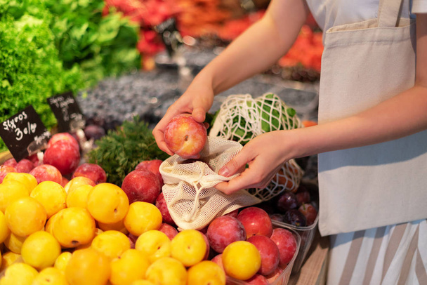 Zero waste, plastic free concept. Sustainable lifestyle. Woman chooses fruits and vegetables at farmers market. Reusable cotton and mesh eco bags for shopping. - 写真・画像