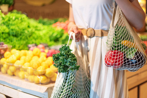Girl holding mesh shopping bag and cotton shopper with vegetables without plastic bags, wooden background. Zero waste, plastic free concept. Sustainable lifestyle. Banner. - 写真・画像