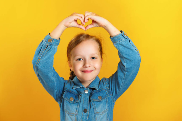 Cheerful girl in a denim shirt on a yellow background. The child shows a heart from fingers above his head and looks at the camera - Photo, image