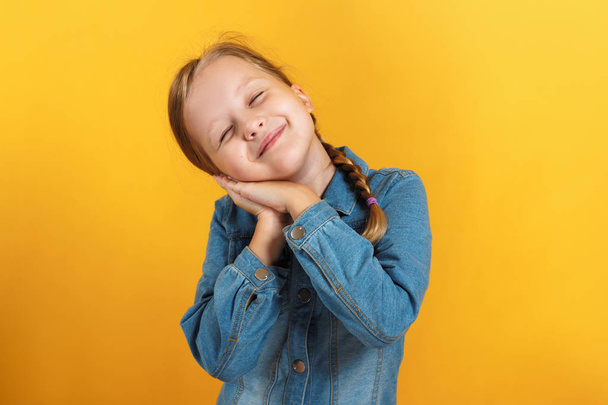 Little girl in a denim shirt on a yellow background. The child closed her eyes and laid her head on folded palms - Photo, Image