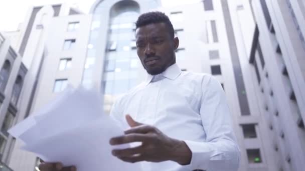 Bottom view of handsome African American man in white shirt reading documents and then throwing them up standing on city street. Male businessman or manager tired and stressed - Filmagem, Vídeo