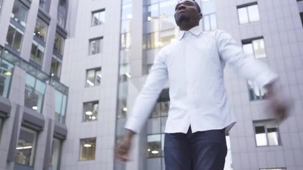 Bottom view of happy African American man in white shirt dancing in front of modern business center on the city street. Male businessman or manager got great news. - Video