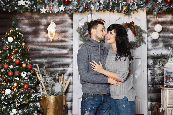 Beautiful couple of lovers, stylish handsome man hugging with beautiful brunette young woman in a trendy grey sweatshirt and posing near the Christmas tree at the interior decorated for New year - 写真・画像