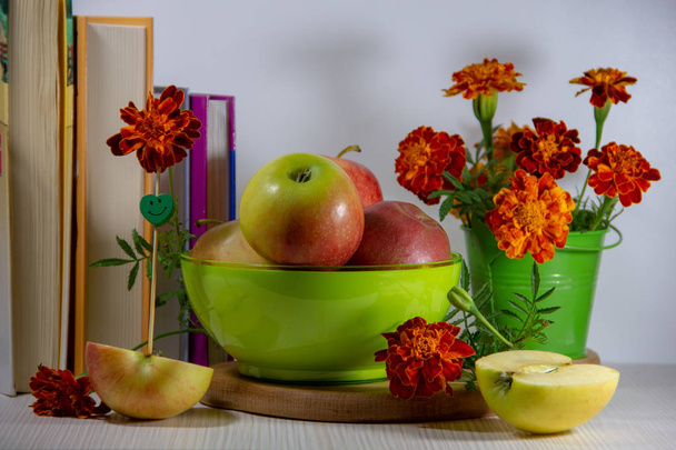 Still life of ripe apples in a green plate, a bouquet of marigolds in a small decorative bucket, books, apple halves with a skewer in the form of an smile on a white background. - Foto, imagen