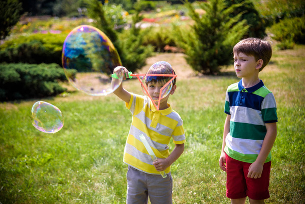 Мальчик надувает мыльные пузыри, в то время как возбужденный ребенок наслаждается пузырьками. Счастливый подросток и его брат в парке наслаждаются созданием мыльных пузырей. Концепция счастливого детства - Фото, изображение