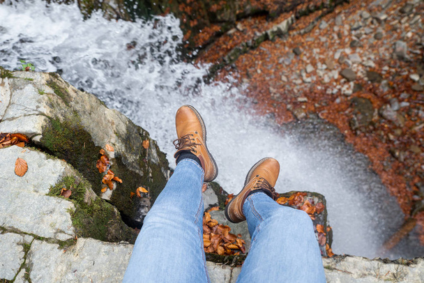 nogi mężczyzna w niebieskie dżinsy i brązowe buty siedzi na krawędzi patrząc na wodospad - Zdjęcie, obraz