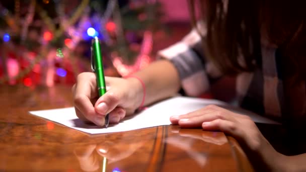 Teen κορίτσι κάθεται σε ένα τραπέζι κοντά σε ένα διακοσμημένο χριστουγεννιάτικο δέντρο και γράφει ένα χριστουγεννιάτικο γράμμα στον Άγιο Βασίλη - Πλάνα, βίντεο