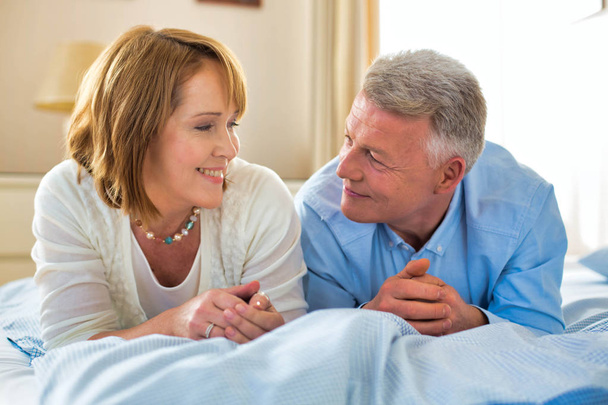 Χαμογελώντας ώριμο ζευγάρι κοιτάζοντας ο ένας τον άλλο, ενώ βρίσκεται στο κρεβάτι στο σπίτι - Φωτογραφία, εικόνα