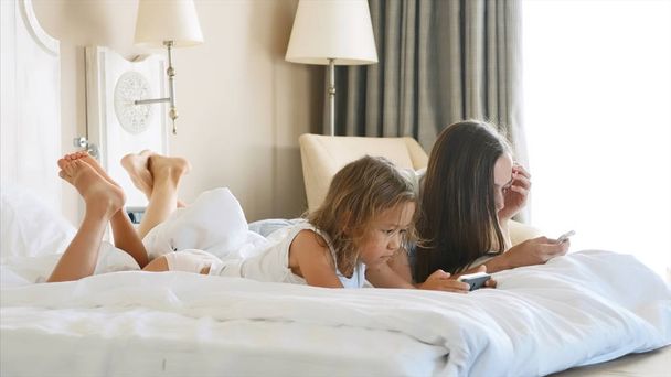 Young mother with little cute daughter are using gadgets lying on a white bed. - Foto, Bild