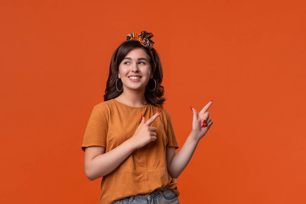 Pretty brunette woman in a t-shirt and beautiful headband pointing with her fingers at the rights side isolated over orange background. Place for ad. - Photo, Image