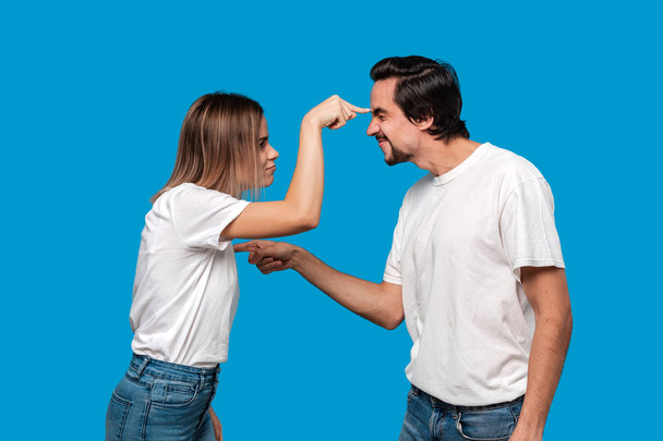 Couple of a young blond woman and brunet bearded man with mustaches in white t-shirts and blue jeans quarreling standing isolated over blue background. - Photo, Image