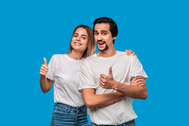 Happy couple of a young blond woman and brunet bearded man with mustaches in white t-shirts and blue jeans posing isolated over blue background. Concept of an ideal couple. Models holding thumbs up - Photo, Image